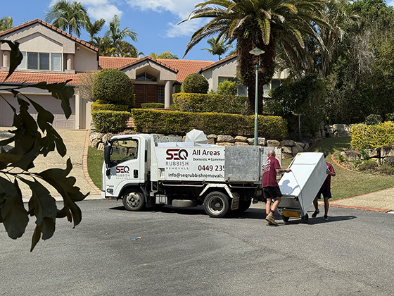 Rubbish removalists with rubbish truck loading old fridge into back for disposal